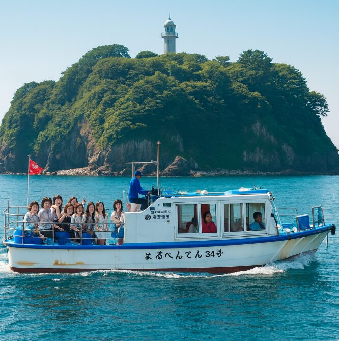 晴れた日に、青い海の上を、多くの乗客を乗せて航行する江の島べんてん丸の遠景。背景には雄大な江の島の緑の丘と切り立った崖が見える